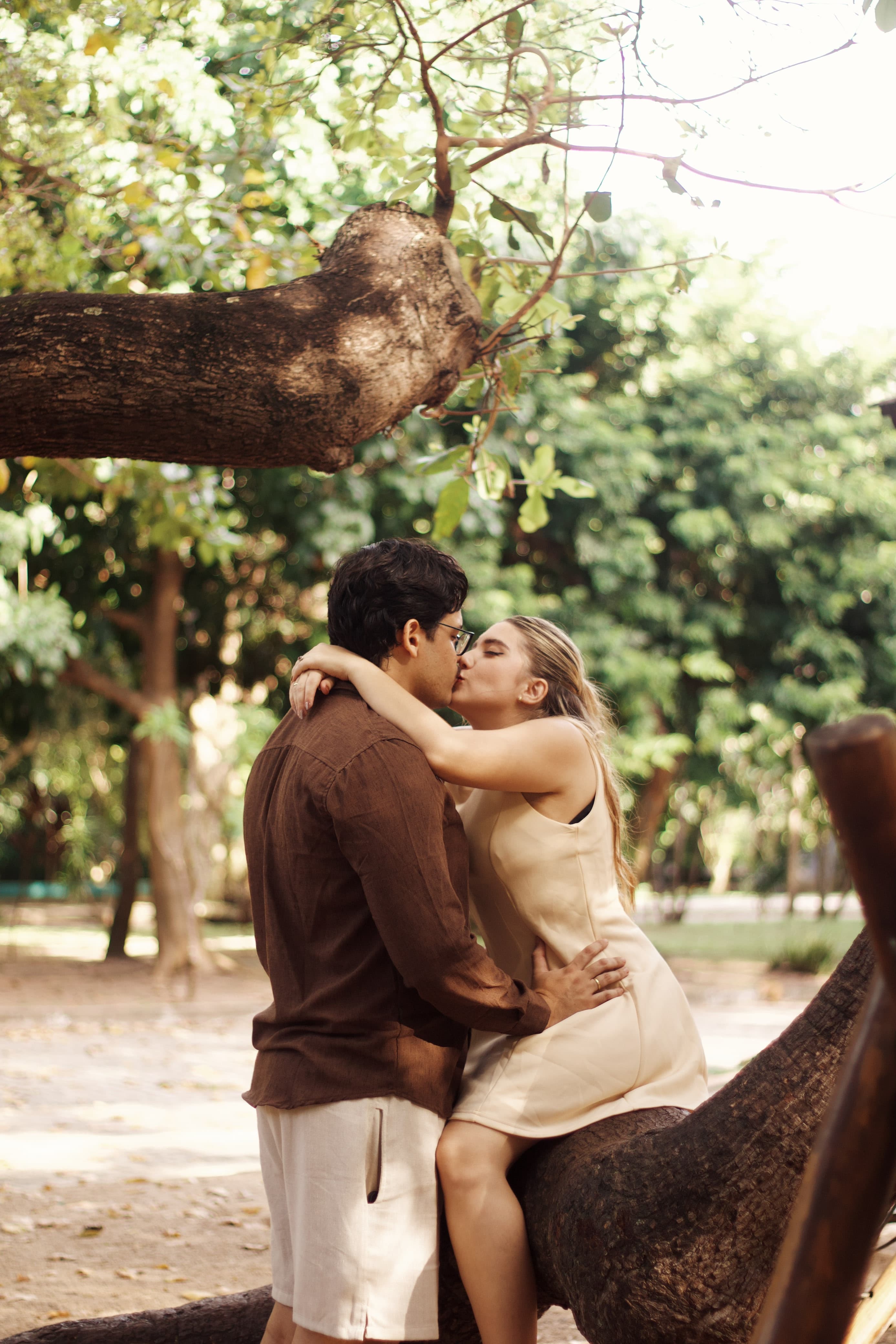 Gabrielle e Pedro se beijam junto a uma árvore, em um jardim arborizado